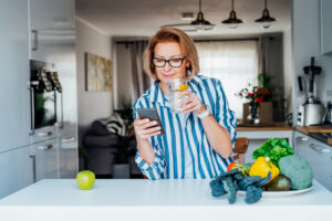 A middle-aged woman in her kitchen, drinking a glass of water while scrolling on her phone.