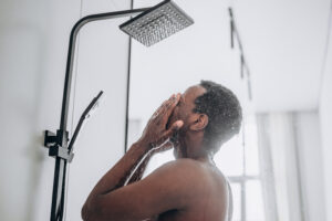 Relaxed young African-American man takes shower standing under hot water jets i