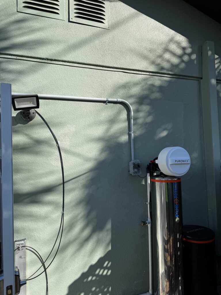 Puronics water softener installed outside a St. Pete Beach home with the plumbing above ground and the electrical outlet mounted high on the wall in conduit for flood protection