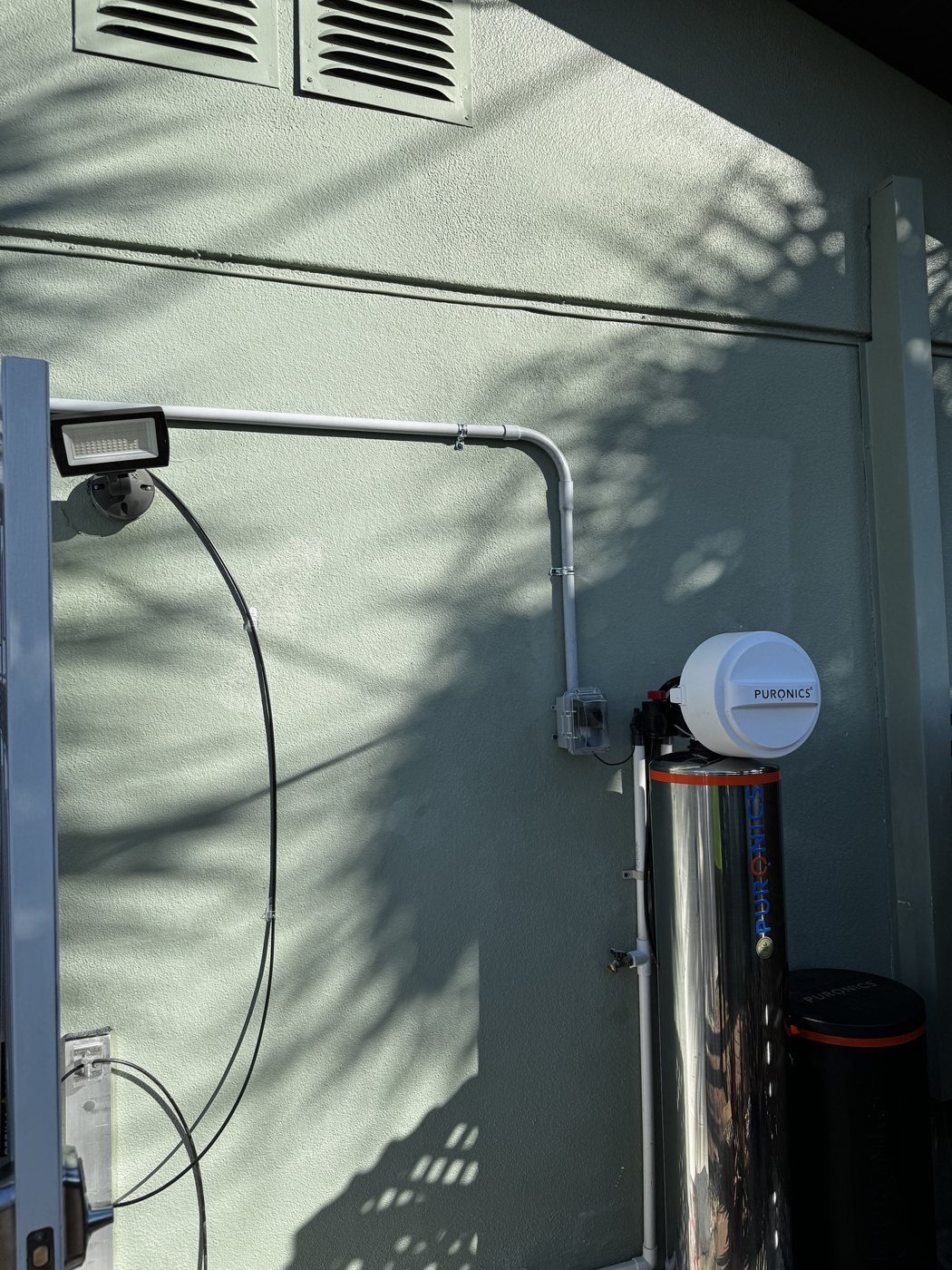 Puronics water softener installed outdoors against a St. Petersburg house, with plumbing run above ground and the electrical outlet mounted high on the wall