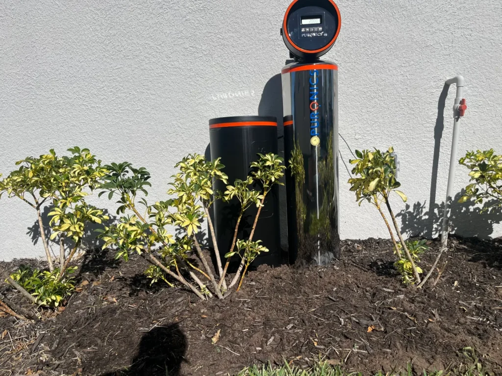 Puronics whole-home water softener and brine tank installed in a landscaped bed beside a home by Hydrology Water Solutions